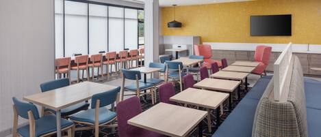 A modern restaurant with multiple tables and chairs, a large window, and a flat-screen TV mounted on the wall.