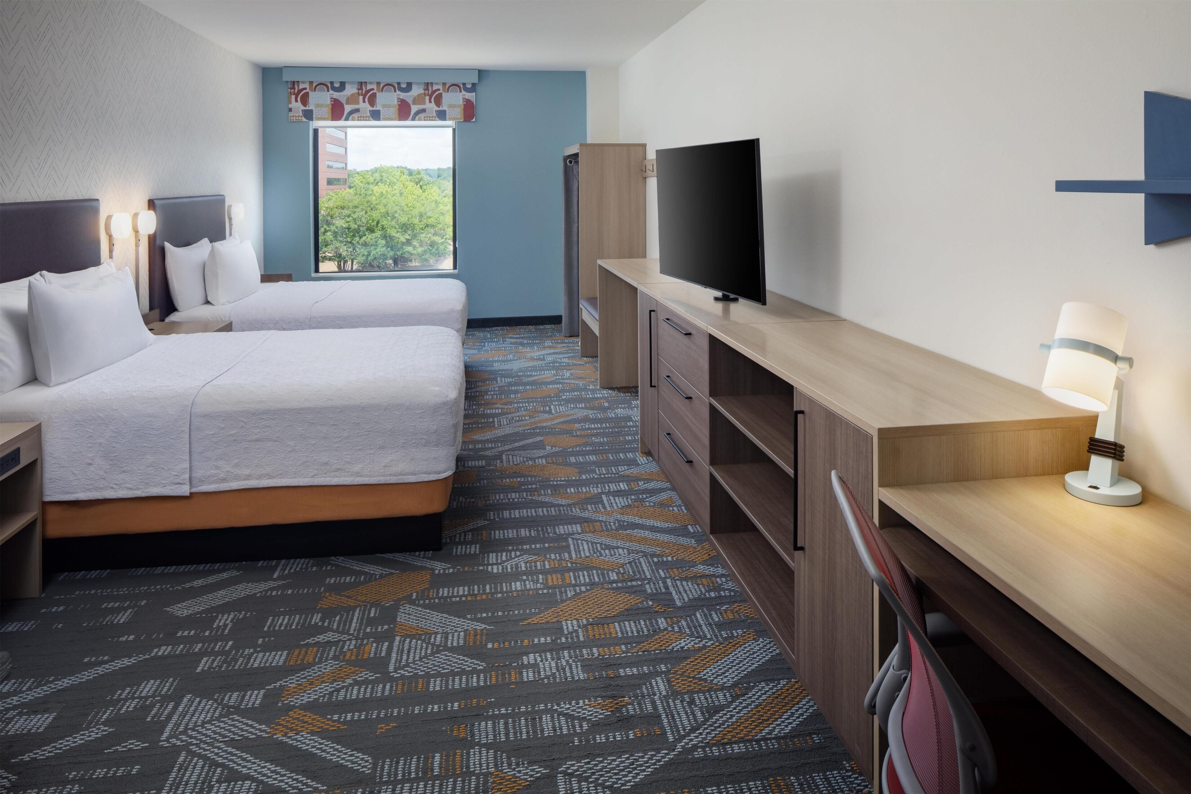 A hotel room with two beds, a flat-screen TV, a desk with a lamp, and a patterned carpet.