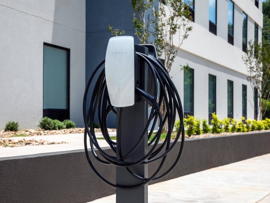 A Tesla charging station with multiple cables connected to it, set against a modern building facade with windows and greenery.