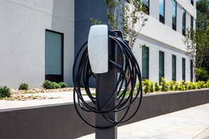 A Tesla charging station with multiple cables connected to it, set against a modern building facade with windows and greenery. - Home2 Suites By Hilton Tyler (Tyler)