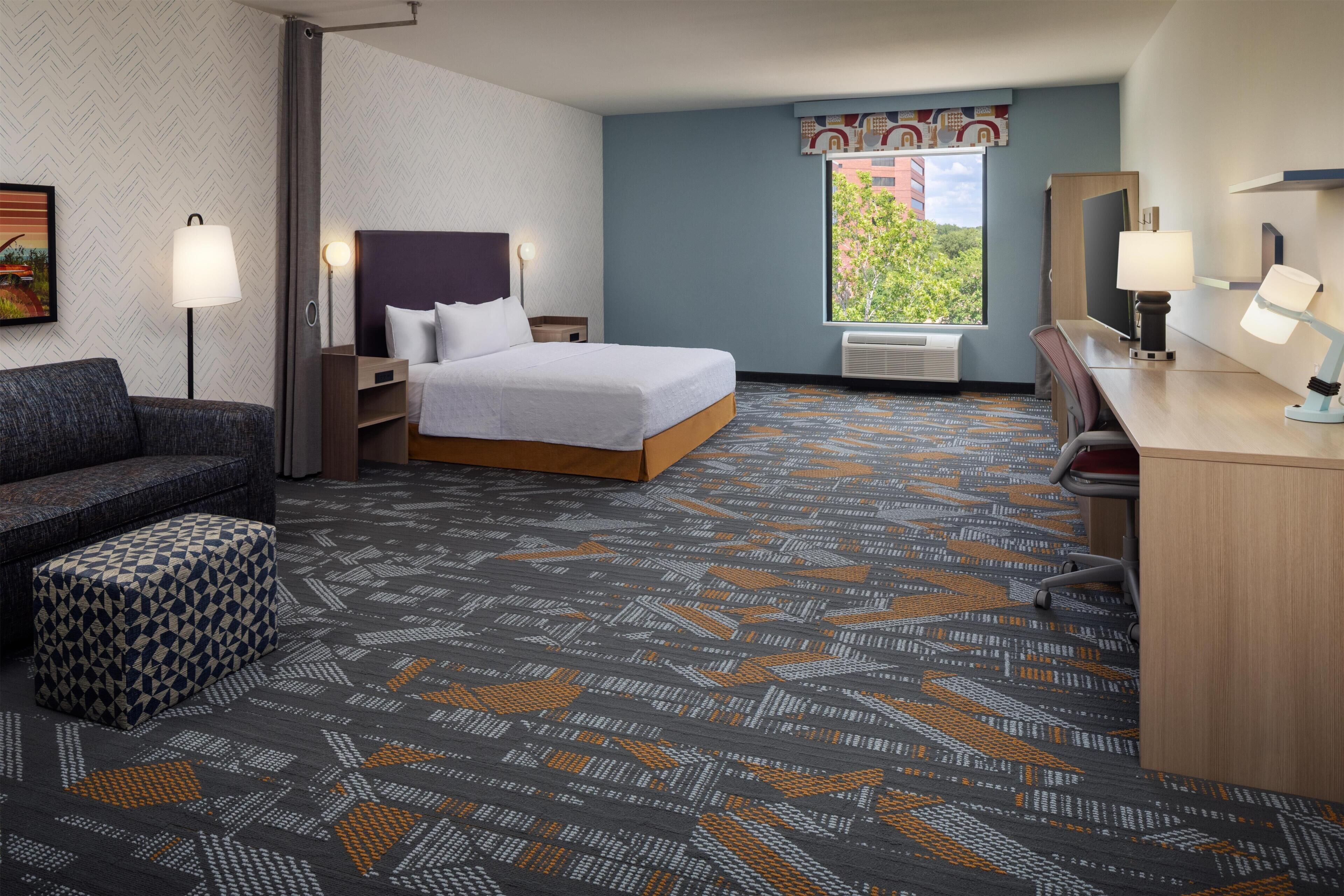 Una habitación de hotel con cama, escritorio, silla, sofá y alfombra estampada.