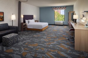 A hotel room with a bed, a desk, a chair, a sofa, and a patterned carpet. - Home2 Suites By Hilton Tyler (Tyler)