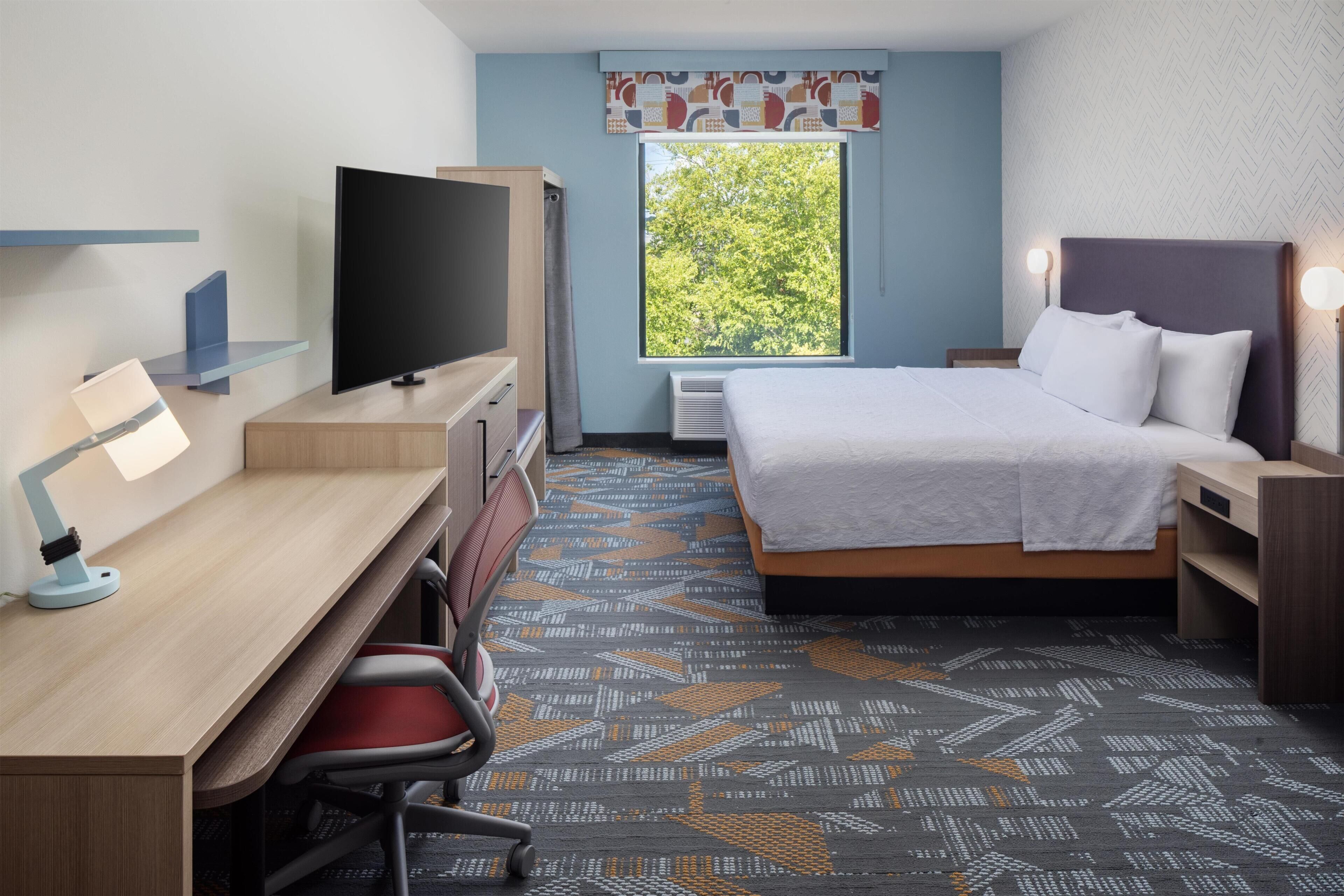 A hotel room with a bed, desk, chair, television, and a window with a view of trees.