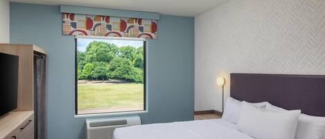 Una habitación de hotel con cama, televisor, ventana con vista a árboles y alfombra estampada.