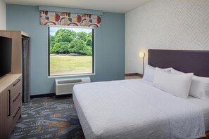 A hotel room with a bed, a television, a window with a view of trees, and a patterned carpet. - Home2 Suites By Hilton Tyler (Tyler)