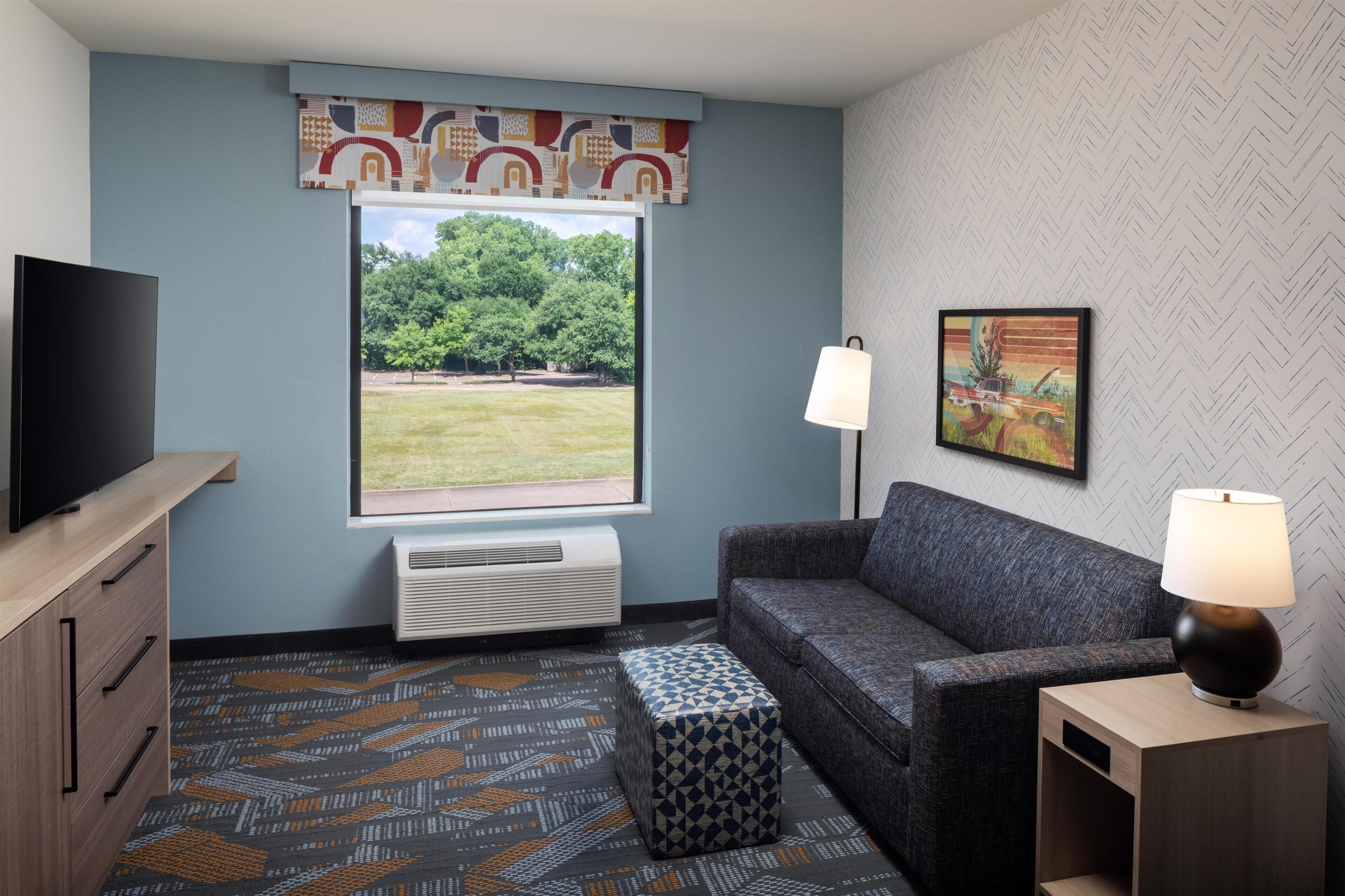 A hotel room with a large window, a flat-screen TV, a sofa, a patterned rug, and a framed picture on the wall.