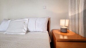 Un letto rifatto con biancheria e cuscini bianchi, un comodino in legno con una lampada, e una parete semplice con un interruttore.