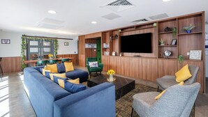 Eine moderne Hotelhalle mit einem blauen Sofa, einem grünen Sessel und einem grauen Sessel. An der Wand ist ein Flachbildfernseher befestigt, es gibt eine Rezeption und einen Essbereich mit grüner Wand und einem Fenster.