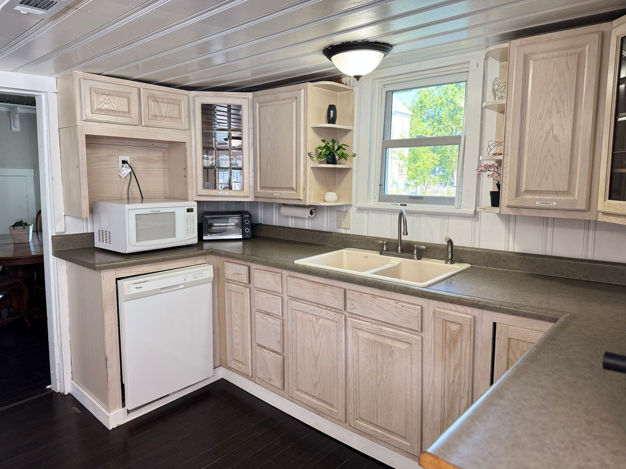 A kitchen with wooden cabinets, a built-in microwave, a dishwasher, and a window with a view of greenery.