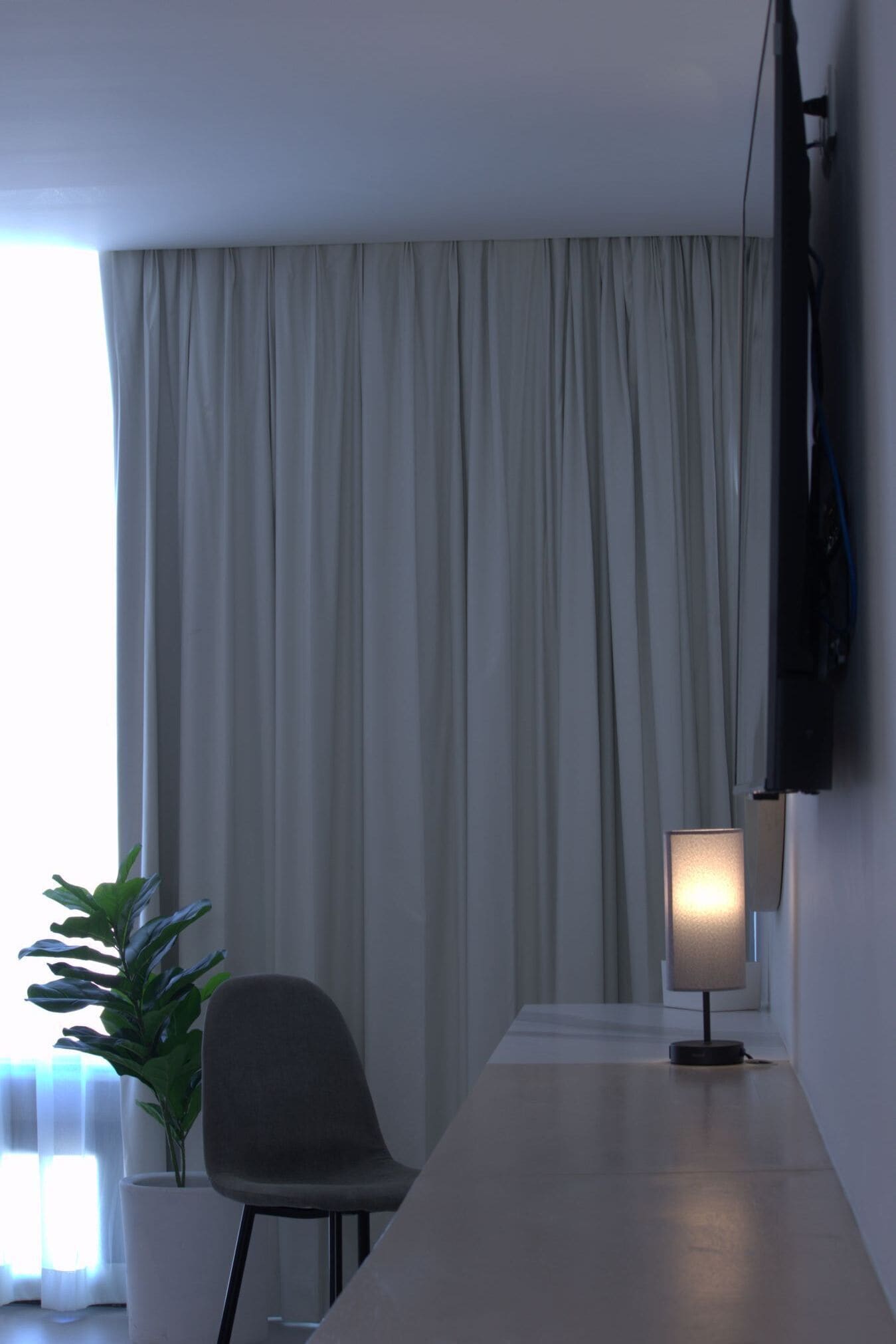 Una habitación moderna con un ventanal grande, un televisor de pantalla plana montado en la pared, un escritorio con lámpara y una silla.