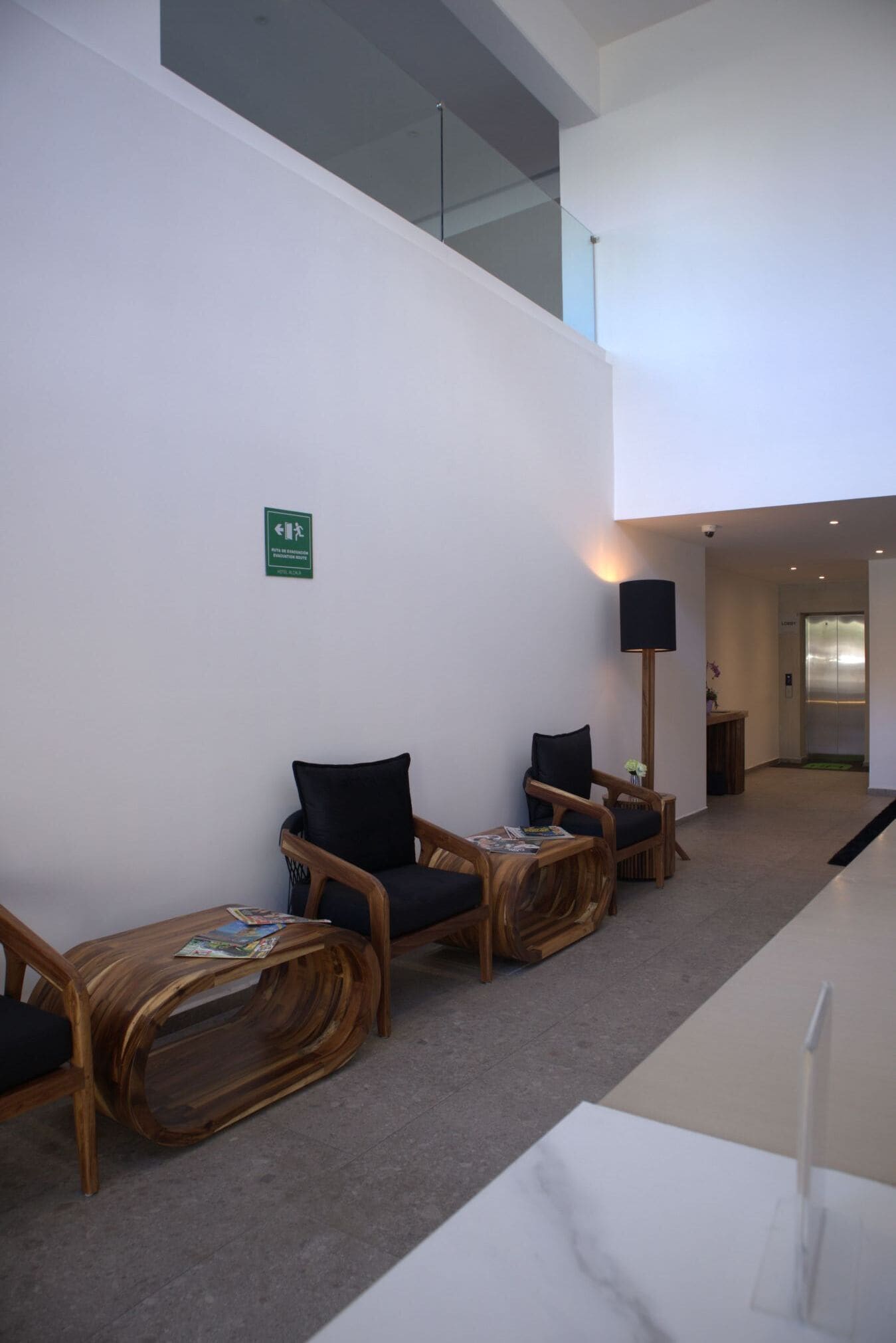 Un área de espera moderna con sillas de madera, una pequeña mesa con revistas y un letrero de salida de emergencia verde en la pared.