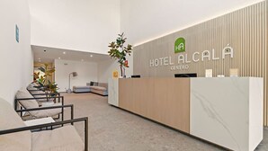 A modern hotel reception area with a wooden reception desk, a seating area with sofas, and a plant. - Hotel Alcalà Centro (Guadalajara)