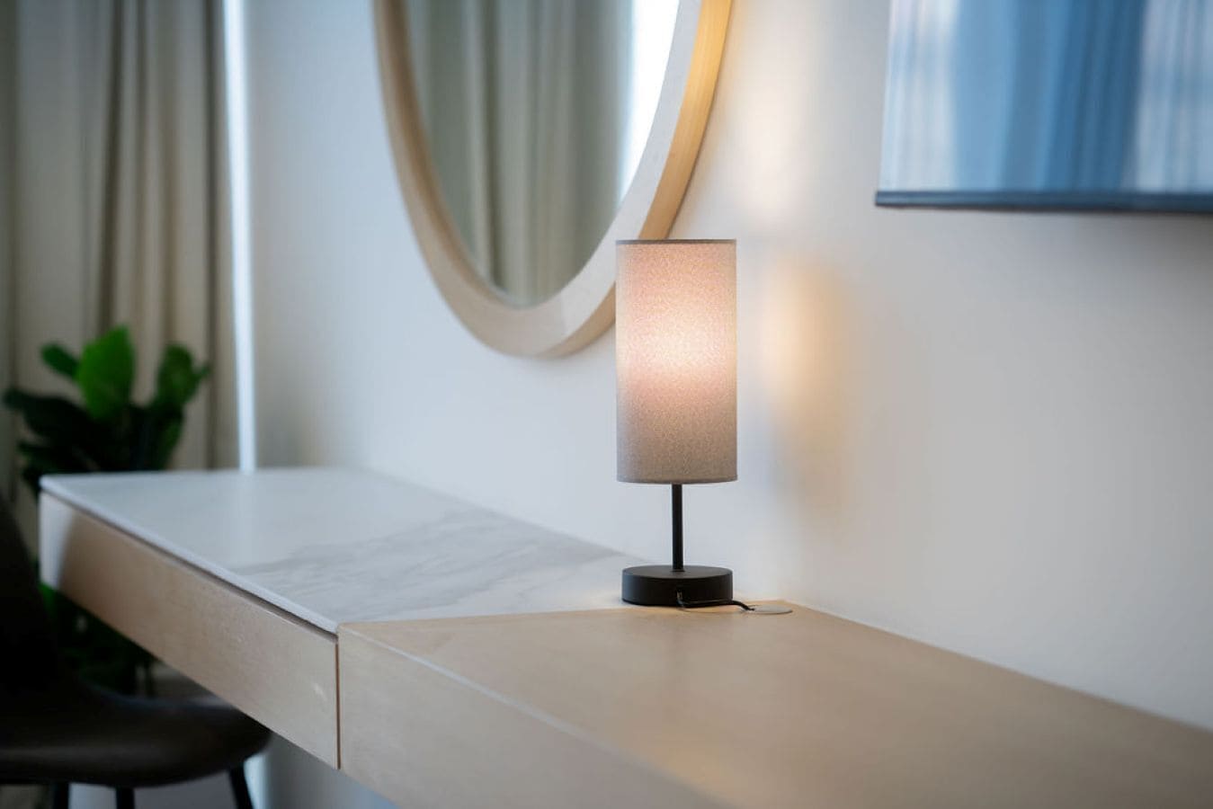 Una lámpara de escritorio moderna con una pantalla cilíndrica, sobre un escritorio de color claro en una habitación con un espejo grande y una planta en maceta.