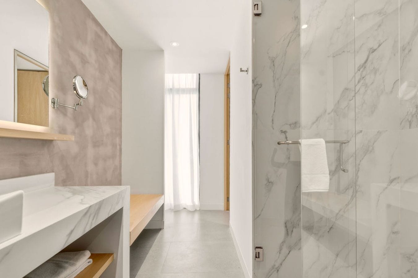 A modern bathroom with a marble shower wall, a wooden vanity, and a mirror with a round frame.