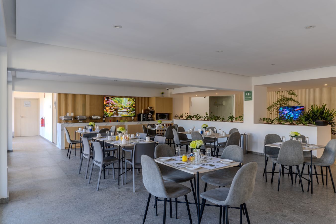 Un espace repas moderne, doté de plusieurs tables et chaises, un coin cuisine équipé d’appareils électroménagers et un écran de télévision diffusant des images de la nature.