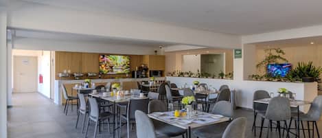 Un espace repas moderne, doté de plusieurs tables et chaises, un coin cuisine équipé d’appareils électroménagers et un écran de télévision diffusant des images de la nature.