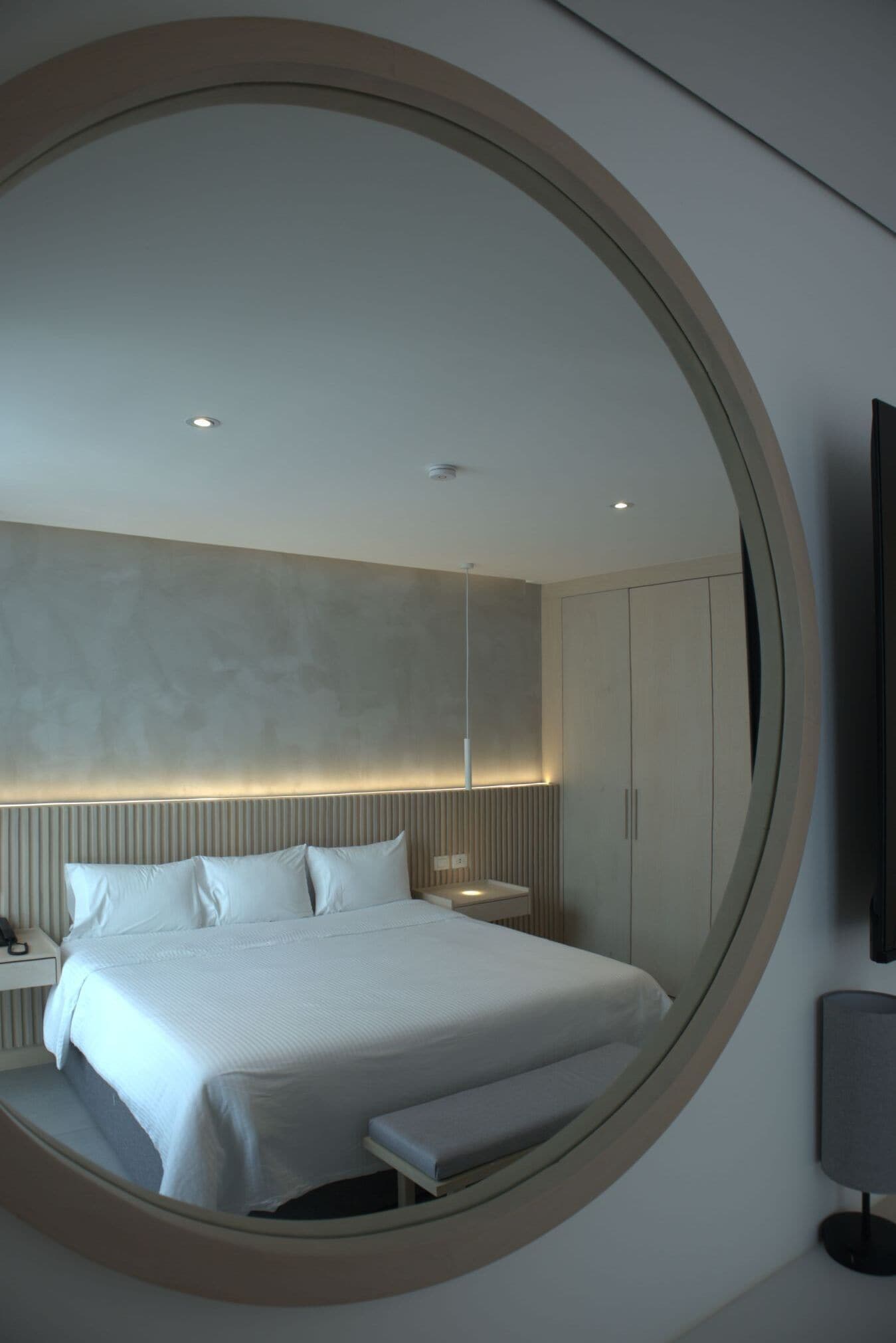 A modern bedroom with a large circular mirror, a bed with white linens, a bedside table, and a wardrobe.
