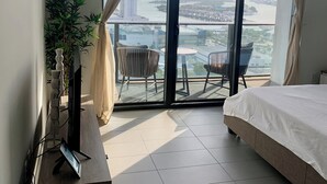 A modern hotel room with a large bed, a TV on a wooden stand, a potted plant, and a balcony with a view of a cityscape and river.