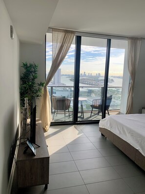 A modern hotel room with a large bed, a TV on a wooden stand, a potted plant, and a balcony with a view of a cityscape and river.