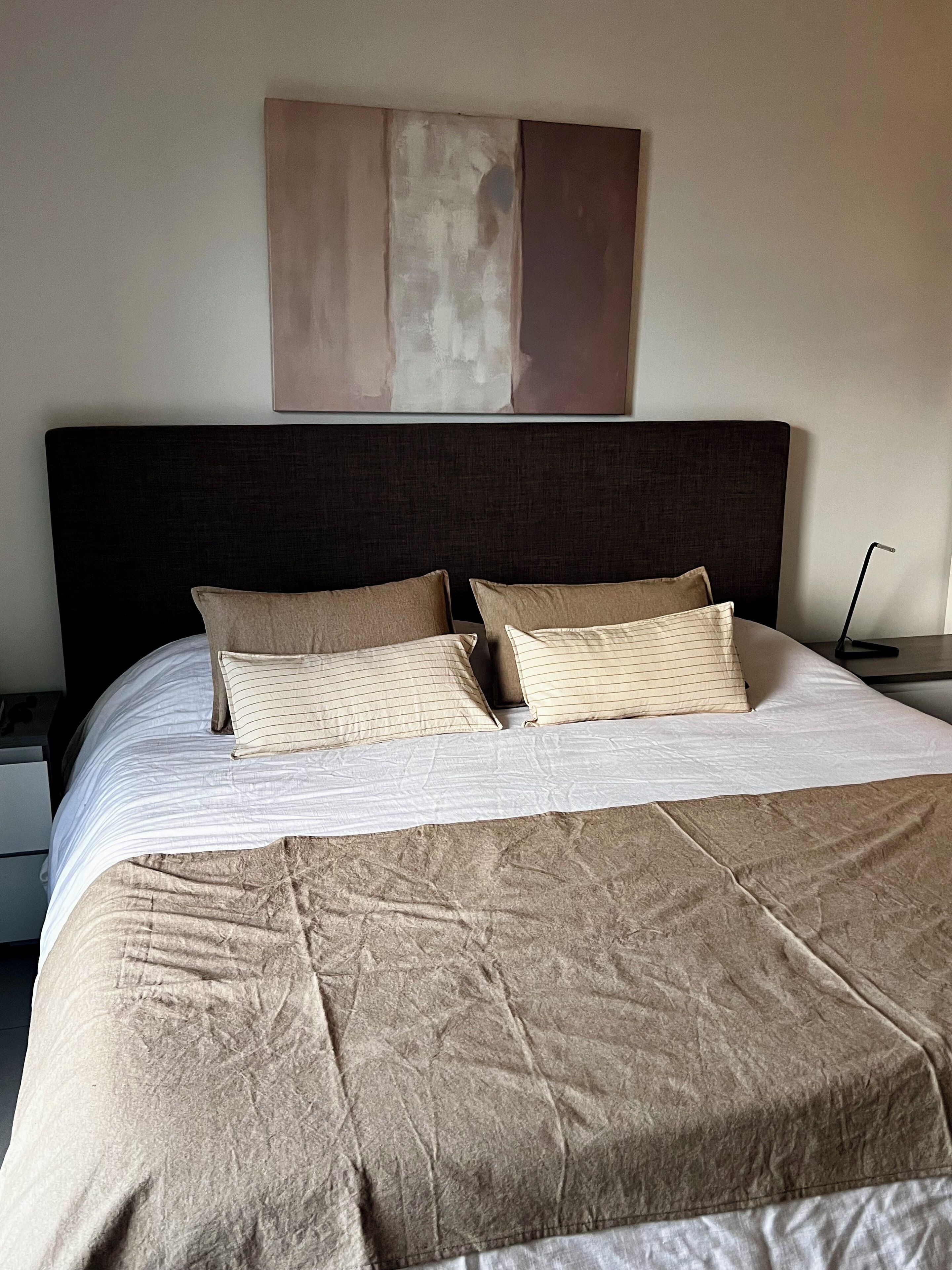 A neatly made bed with a dark headboard and two pillows, a plain wall with a large abstract painting, and a bedside table with a lamp.