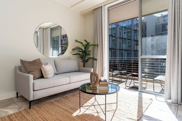 A modern living room with a grey sofa, a round mirror, a small round table with books, a potted plant, and a balcony with a view of buildings.