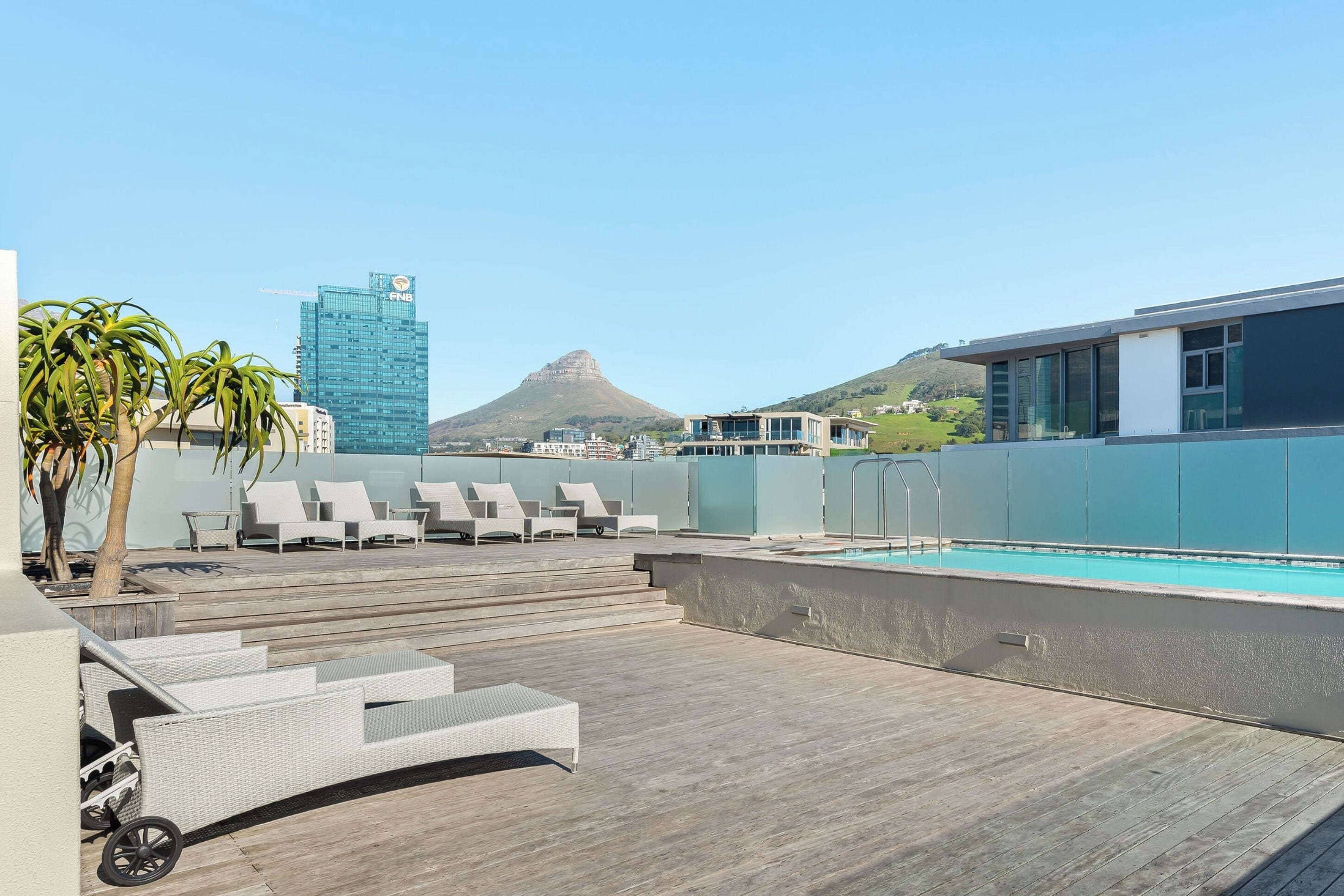 A rooftop pool area with lounge chairs, a pool, and a view of a mountain and a building.
