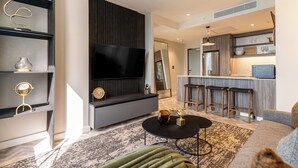 A modern living room with a flat-screen TV, a coffee table, and a sofa. There is a kitchen area with a bar and stools.