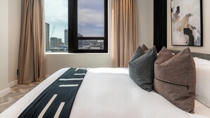 A hotel room with a large bed, beige curtains, a city view, and a modern abstract painting.