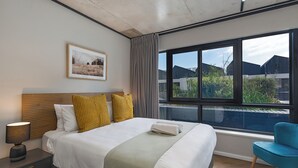 A modern hotel room with a large bed, a nightstand, a lamp, a framed picture on the wall, a window with a view of buildings, and a blue chair. - Cosy and Charming Bridgewater One Apartment | Wifi (Cape Town)