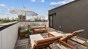 Eine Dachterrasse mit einer Couch, Liegestühlen, einem Couchtisch und einem gestreiften Sonnenschirm.