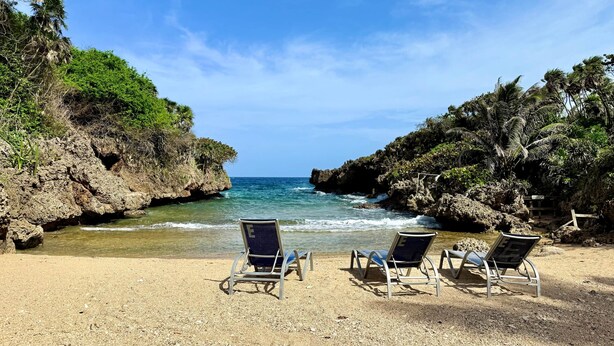 Una playa con acantilados rocosos y agua azul cristalina. Se observan tres sillas de playa vacías orientadas al mar.