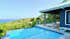 Una piscina con vista al océano, rodeada de vegetación y una terraza de madera con sillas reclinables.