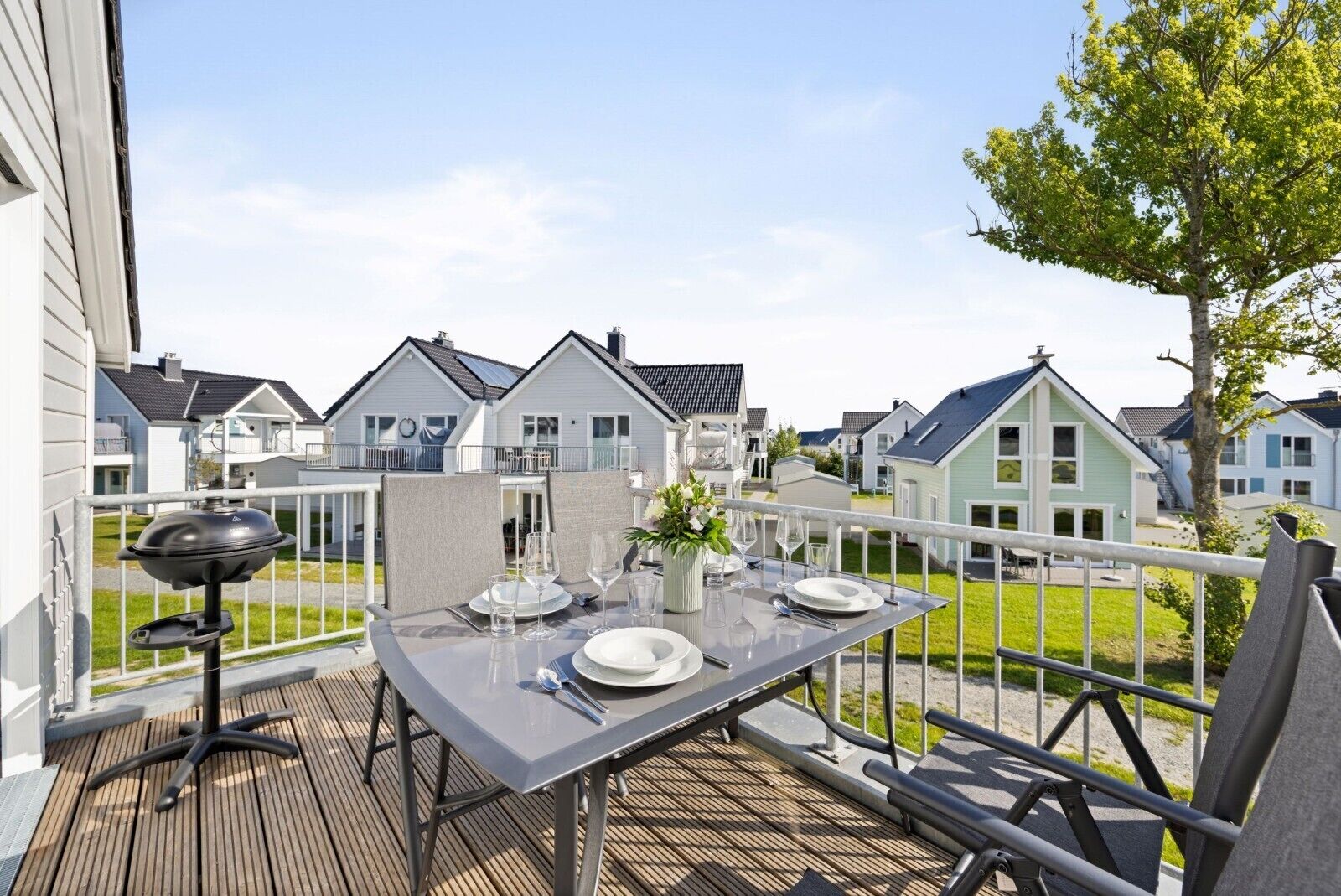 An outdoor dining area with a table set for two, a barbecue grill, and a view of neighboring houses.