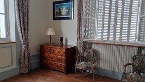 A room with a wooden dresser, a chair, a lamp, a framed picture, and a window with blinds. - Guest room on 1st floor of château (Hinacourt)