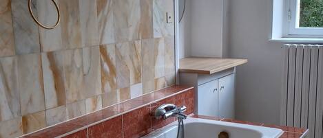 A bathroom with a white bathtub, red and beige marble tiles, a gold towel ring, and a window with a view of greenery.
