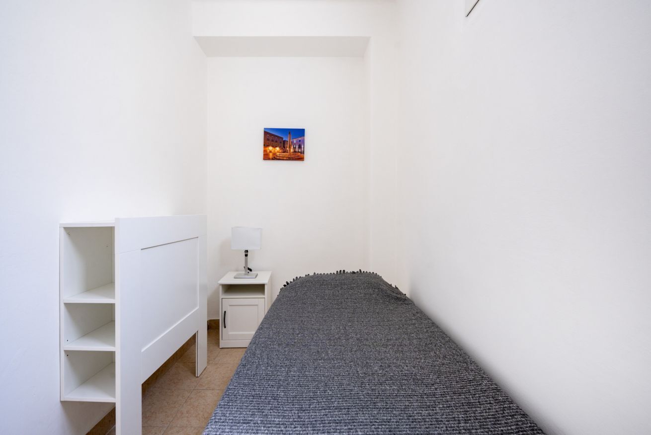 A small, neatly arranged bedroom with a bed, a nightstand, a lamp, a shelf unit, and a framed picture on the wall.