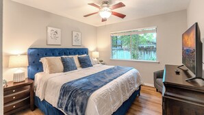 Une chambre avec un grand lit, un ventilateur de plafond, une fenêtre donnant sur l’extérieur et un téléviseur posé sur un meuble.