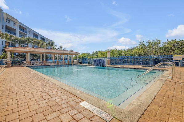 A swimming pool with a tiled deck, a covered seating area, and a building with balconies in the background.