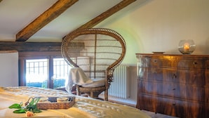 A cozy bedroom with a large bed, a wicker chair, a wooden dresser, and a window with a view. - Foresteria Casaforte Bergum at the gates of Cividale (Friuli-Venezia Giulia)