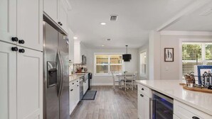 A modern kitchen with white cabinetry, a stainless steel refrigerator, and a dining area with a table and chairs. - Gator Shack, Luxury Chic! (Gainesville)