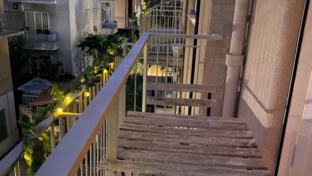 Un banc en bois sur un balcon doté d’une balustrade, offrant une vue sur un quartier résidentiel avec ses immeubles et sa verdure.