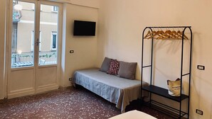 Une petite pièce bien éclairée, avec un lit, un téléviseur fixé au mur, une fenêtre donnant sur des immeubles et un ventilateur de plafond.
