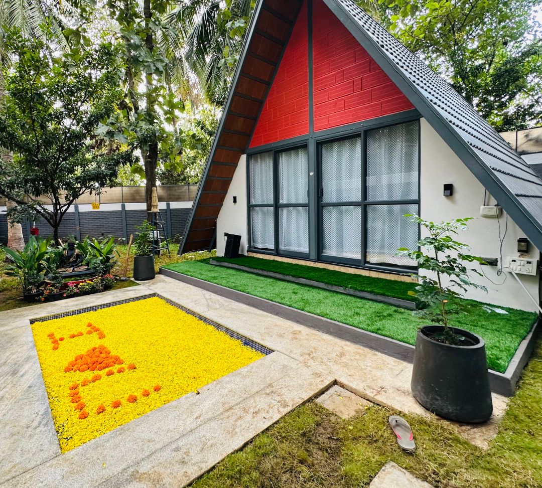 A modern, triangular-roofed building with a red exterior and large windows, surrounded by lush greenery and a yellow patterned area.
