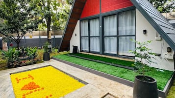 A modern, triangular-roofed building with a red exterior and large windows, surrounded by lush greenery and a yellow patterned area.