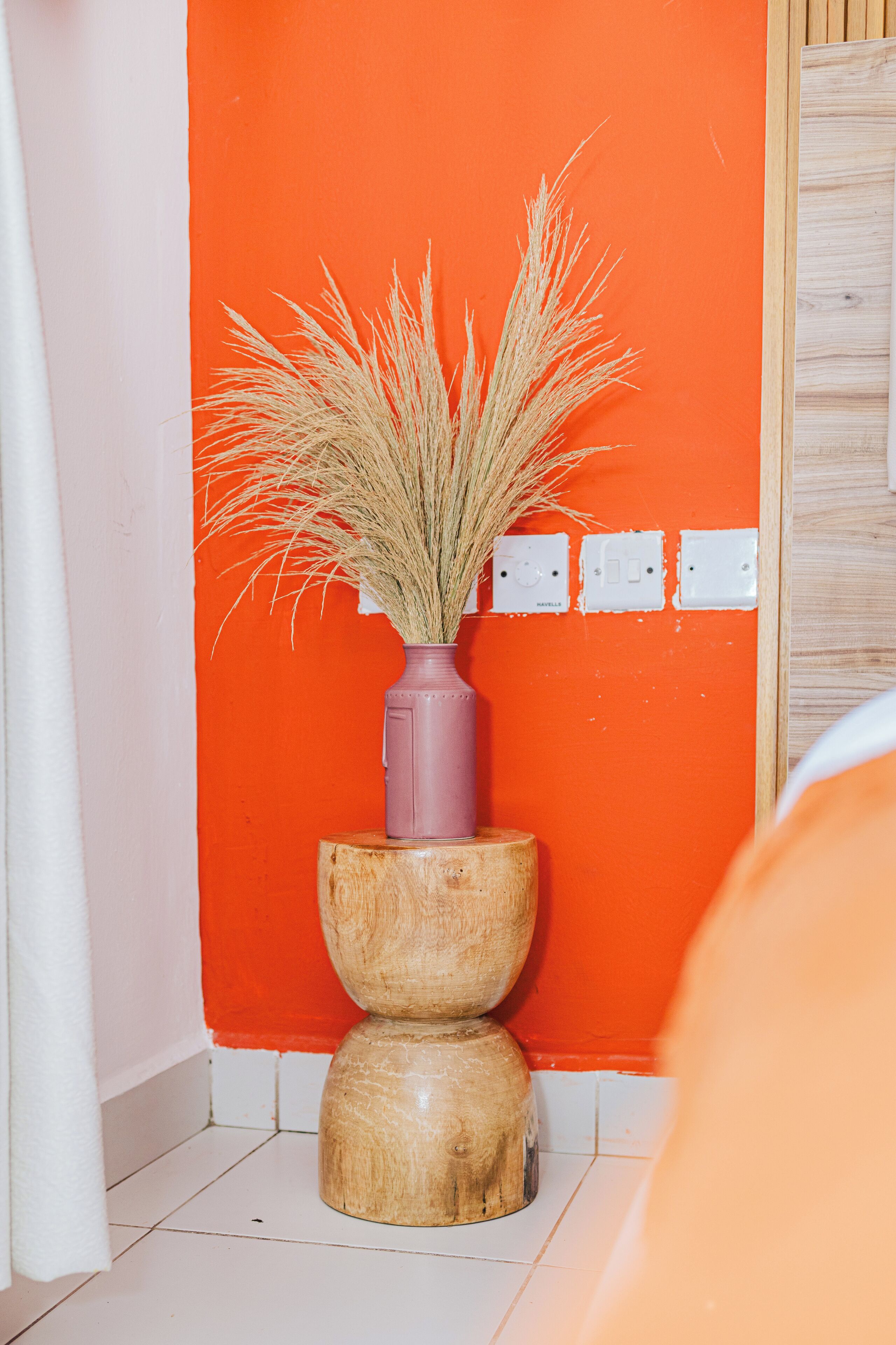 Ein Badezimmer mit einem hölzernen Hocker, einer Vase mit Trockenpflanzen und einer leuchtend orangefarbenen Wand.