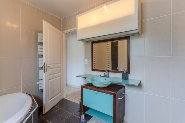 Une salle de bain avec une baignoire, un lavabo en verre, un meuble-lavabo en bois, un miroir et une porte.