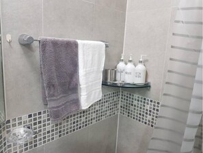 Un baño con un perchero que sostiene una toalla morada y blanca, un estante de vidrio con artículos de aseo y una zona de ducha con paredes de azulejos.