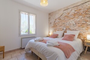 A bedroom with a brick wall, a bed with white bedding, a window with curtains, and a bedside table with a lamp. - Domaine Plan des Devens – Peaceful 4-Star Retreat with Heated Pool (Roussillon)