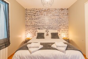 A bedroom with a brick-patterned wall, a bed with white bedding, two bedside tables with lamps, and a window with curtains. - Domaine Plan des Devens – Peaceful 4-Star Retreat with Private Terrace (Roussillon)