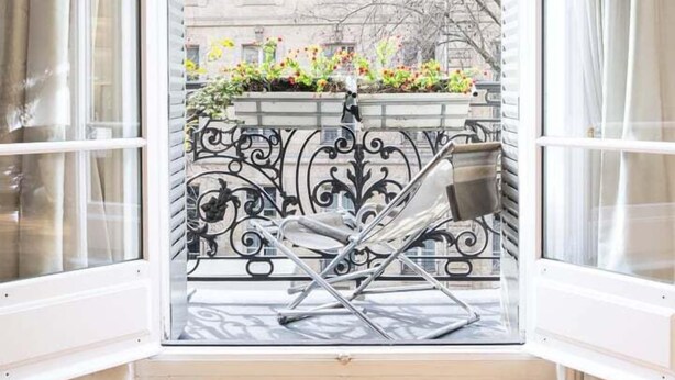 A room with a large window, white curtains, and a view of a building with balconies and flowers.